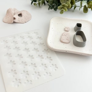 May include: A white silicone mold with multiple flower-shaped cavities, a pink silicone mold with a flower design, and two gray silicone cutters, one shaped like a flower and the other shaped like a rectangle.