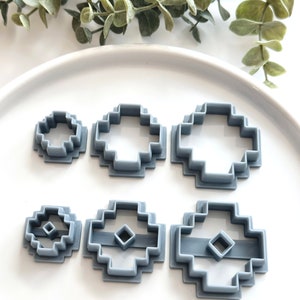 Puede incluir: Conjunto de seis cortadores de galletas de plástico gris en varios tamaños y formas. Los cortadores tienen forma de círculo, cuadrado y cruz con un agujero cuadrado en el centro.