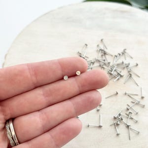 May include: Two small, round, gold-colored stud earrings. The earrings are held in a hand, with a blurred background of small, silver-colored posts. The earrings have a simple, minimalist design.