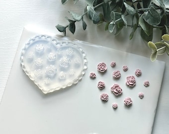 May include: A heart-shaped silicone mold filled with rose designs, alongside several small, light pink rose embellishments. The mold has a scalloped edge. Greenery is in the background.