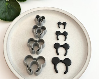 May include: A selection of grey and black Mickey Mouse ear-shaped cookie cutters of different sizes, displayed on a white plate. The cutters are made of plastic and are for baking. The image is well-lit, highlighting the details of the cutters.