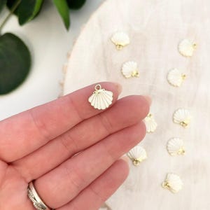 May include: A collection of small, white seashell charms with gold-colored accents. The charms are held in a hand, and several others are scattered on a light-colored wooden surface. These charms are ideal for jewelry making or craft projects.
