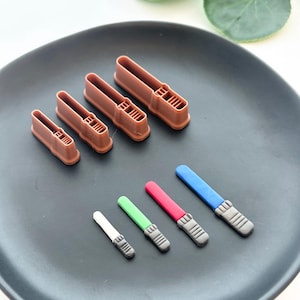 May include: Brown plastic lightsaber-shaped cookie cutters in various sizes, with miniature lightsaber-shaped sweets in white, green, red, and blue. The items are on a dark grey plate.