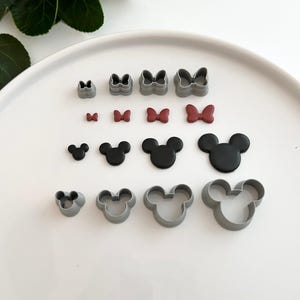 Peut inclure: Un ensemble de 14 emporte-pièces en plastique gris en forme de nœud et de tête de Mickey. Les emporte-pièces sont disposés sur une surface blanche. Il y a quatre tailles de chaque forme.