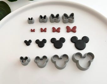 May include: A set of 14 grey plastic cookie cutters in the shapes of a bow and Mickey Mouse head. The cutters are arranged on a white surface. There are four sizes of each shape.
