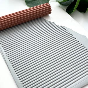 Könnte beinhalten: Ein brauner, geriffelter Nudelholz liegt auf einem grauen, strukturierten Blatt mit parallelen Linien. Das Blatt liegt auf einer weißen Oberfläche, mit grünen Blättern im Hintergrund. Die Walze wird wahrscheinlich zum Erstellen von strukturierten Mustern verwendet.