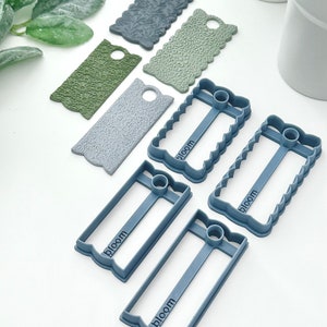 May include: A set of six blue plastic cookie cutters with a scalloped edge and the word "bloom" embossed on each. The cutters are in the shape of a rectangle with a hole in the center.