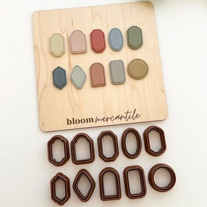 May include: A wooden board with various pastel-colored shapes and brown cookie cutters. The board has the text "bloom mercantile" printed on it. The shapes are in shades of pink, blue, green, and tan. The cookie cutters are in various geometric shapes.