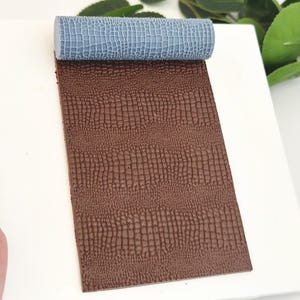 以下が含まれることがあります： ワニの皮の模様がついた茶色のテクスチャ加工されたシリコン製の麺棒。麺棒は白い表面の上に置かれています。