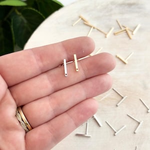 May include: Two pairs of minimalist bar earrings, one pair in silver and one pair in gold. The earrings are shown on a hand, with the silver pair on the left and the gold pair on the right.