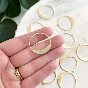 Puede incluir: Pendiente de círculo de metal dorado con una superficie texturizada y un semicírculo recortado. El pendiente se muestra sobre una superficie blanca con otros pendientes similares.