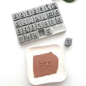 Pode incluir: Um conjunto de carimbos de alfabeto cinza com o texto "bloom mercantile" na lateral. Um pequeno pedaço de argila em um prato branco salpicado é carimbado com a palavra "hey".