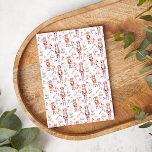 May include: A rectangular sheet of paper with a repeating pattern of red and white nutcrackers, candy canes, and floral elements. The design is set against a white background. The paper is placed on a wooden tray.