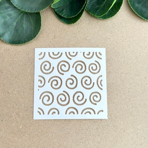 May include: White stencil with a repeating pattern of swirls. The stencil is on a brown background.