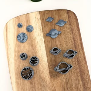May include: A set of nine gray plastic cookie cutters shaped like planets with rings. The cutters are arranged on a wooden cutting board.