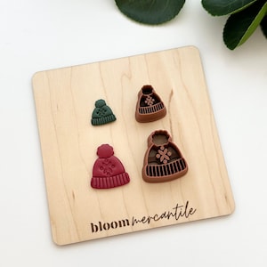 Peut inclure: Quatre petits emporte-pièces en forme de chapeau, verts, marron et rouge-bordeaux, disposés sur une planche en bois clair. La planche porte l'inscription "bloom mercantile" en noir. Les découpoirs ont des détails de flocons de neige et de côtes.