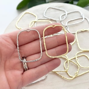 May include: A hand holding two metal jewelry findings. One is silver, the other gold. Both are rectangular with a textured finish and a small loop at the top. Several other gold and silver findings are scattered on a light-colored surface.
