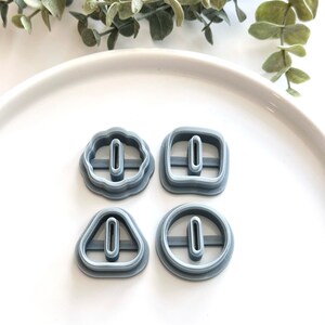May include: Four gray plastic cookie cutters in different shapes: a flower, a square, a triangle, and a circle. Each cutter has a small rectangular hole in the center.