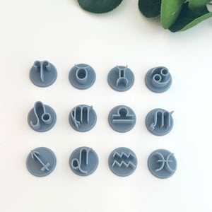Op de afbeelding: Een set van 12 grijze plastic koekjesvormen in de vorm van sterrenbeelden. De vormen zijn gerangschikt in drie rijen van vier. De tekens zijn: Ram, Stier, Tweelingen, Kreeft, Leeuw, Maagd, Weegschaal, Schorpioen, Boogschutter, Steenbok, Waterman en Vissen.