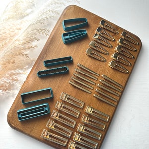 May include: A wooden cutting board displays various hair accessories. Teal-colored plastic hair clip molds are arranged alongside gold-toned metal barrettes and hair clips. The items are set against a white background with dried ornamental grass.