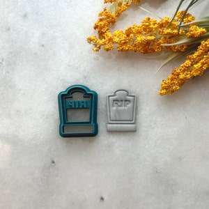 May include: Two cookie cutters, one teal and one gray, shaped like a tombstone with the word "RIP" on it.