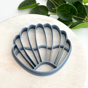 Könnte beinhalten: Ein grauer Plastik-Ausstecher in Form einer Muschel. Der Ausstecher ist muschelförmig mit einem gewellten Rand und einem erhabenen Design.