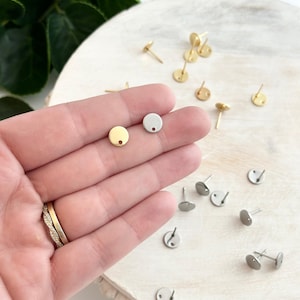 May include: A close-up of two earring blanks, one gold and one silver, with a small hole in the center. The blanks are on a hand, and there are more blanks scattered on a wooden surface.