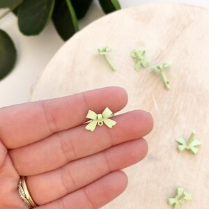 May include: A collection of small, light green bow-shaped charms. One charm is held in a hand, while others are scattered on a light-colored surface. The charms have a delicate, minimalist design, suggesting they are for jewelry or craft projects.