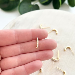 Puede incluir: Aretes de aro dorados con pequeñas perlas blancas. Los aretes se muestran en una mano y sobre una superficie blanca.