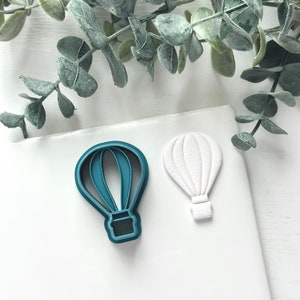 May include: Two hot air balloon cookie cutters, one teal blue and one white. The teal blue cutter has a detailed design with lines and a basket. The white cutter is a simple silhouette.
