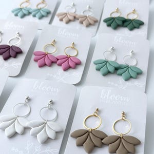 Könnte beinhalten: Sortiment handgefertigter Ohrringe aus Polymer Clay mit goldenen und silbernen Creolen. Die Ohrringe sind in verschiedenen Farben erhältlich, darunter Rosa, Grün, Blau, Braun und Weiß. Die Ohrringe werden auf weißen Karten mit dem aufgedruckten Text "bloom mercantile" präsentiert.