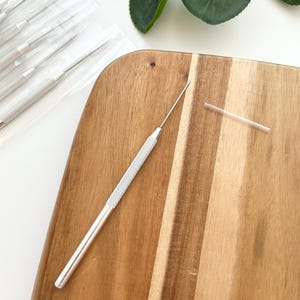 May include: A silver metal weeding tool with a fine, sharp needle point and a textured, knurled grip handle. The tool is accompanied by a small, clear plastic protective cap and is part of a set of similar tools packaged in individual clear sleeves.