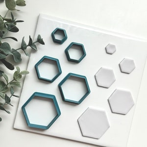 May include: A set of teal hexagon cookie cutters and white hexagon clay shapes on a white surface.