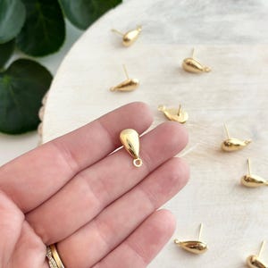 May include: Gold-colored teardrop-shaped stud earrings. The earrings have a smooth, polished finish and a post for insertion. Several earrings are scattered on a light-colored wooden surface, with a hand holding one earring in the foreground.
