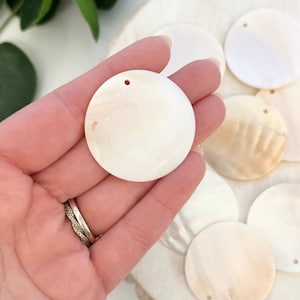 Puede incluir: Una colección de colgantes redondos de concha de color blanquecino con un brillo nacarado. Cada colgante tiene un pequeño agujero cerca de la parte superior. Los colgantes se sostienen en una mano, con varios otros visibles en el fondo, lo que sugiere un tema de artesanía o joyería.