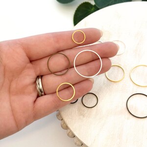 May include: A collection of small, circular metal rings in various colors, including gold, silver, and black. The rings are displayed on a wooden surface, with some held in a hand. The rings vary in size and are suitable for jewelry making or crafting.