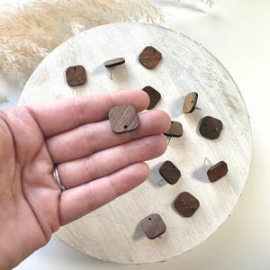 May include: A close-up of a hand holding a set of 10 wooden square earrings with a hole in the center. The earrings are a dark brown color and have a natural wood grain.