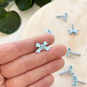 May include: Light blue bow-shaped stud earrings. The earrings are small and delicate, with a bow design. Several pairs of earrings are scattered on a light wooden surface. The earrings are a pastel blue color.
