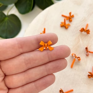 May include: Close-up of small orange bow-shaped embellishments. The bows are scattered on a light-colored surface. The bows are made of a solid material and have a simple, symmetrical design. The bows are a bright, cheerful orange color.