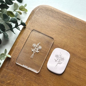 Puede incluir: Un sello de acrílico transparente con un dibujo lineal simple de un ramo de flores. El sello está sobre una superficie de madera y hay una impresión de arcilla rosa del sello al lado. El sello está etiquetado como "Bloom Mercantile".