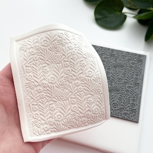May include: A white, floral-patterned silicone mold with a curved edge, held in a hand. A gray, floral-patterned mold and a white surface are also visible. The molds feature a detailed botanical design, including flowers and leaves. The text "BLOOM MERCANTILE" is printed on the side of the white mold.