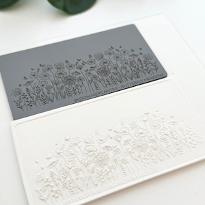 Może przedstawiać: Silikonowa stempelka w kolorze szarym i białym z kwiatowym wzorem. Stempelka przedstawia różne kwiaty i motyle. Tekst "Bloom Mercantile Butterfly Meadow" jest nadrukowany na szarej stempelce.