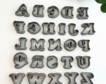 May include: A collection of grey plastic alphabet cookie cutters, arranged in rows. The letters are in a rounded, bold font. Green leaves are visible in the background.
