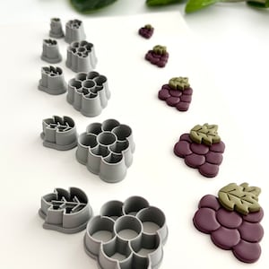 Könnte beinhalten: Ein Set mit 10 grauen Plastik-Ausstechern zum Ausstechen von Trauben- und Blattformen. Die Ausstecher sind in zwei Reihen auf einer weißen Oberfläche angeordnet. Es gibt drei verschiedene Größen von Trauben-Ausstechern und drei verschiedene Größen von Blatt-Ausstechern. Außerdem gibt es vier kleine runde Ausstecher.