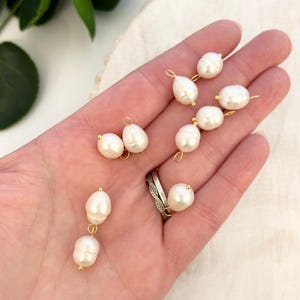Puede incluir: Una colección de perlas blancas en forma de lágrima con bucles y detalles de alambre dorado. Las perlas varían ligeramente en tamaño y forma, dispuestas sobre una superficie clara. La imagen muestra el brillo y el diseño delicado de las perlas.