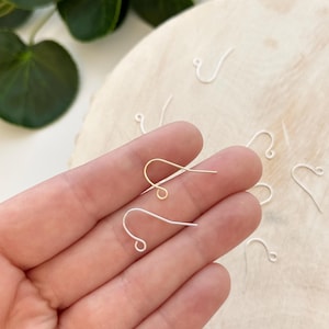 May include: Two earring hooks, one gold and one silver, are held in a hand. The hooks are made of thin metal and have a small loop at the top.