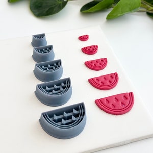 Pode incluir: Um conjunto de sete cortadores de argila polimérica em forma de melancia, cinzentos e um rosa. Os cortadores estão dispostos em uma fila do menor para o maior. Os cortadores são usados para criar biscoitos, fondant ou outras artesanato em forma de melancia.