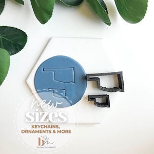 May include: Two black cookie cutters in the shape of the state of Oklahoma. The cutters are on a white surface with a blue circle in the background. The circle has a smaller cookie cutter inside of it. The text "new sizes" is written in black and white on the surface. The text "keychains, ornaments & more" is written in black and white below the text "new sizes".
