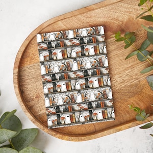 May include: A rectangular notepad with a repeating pattern of white ghosts, orange mushrooms, and brown books on wooden shelves against a gray background. The notepad is placed on a wooden tray with a natural finish.