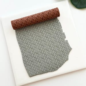 May include: A brown textured rolling pin with a leaf pattern rests on a gray clay sheet, also with a leaf pattern. The clay sheet is on a white square tile. The rolling pin is used for crafts and creating textures.